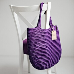 Peut inclure: Un sac fourre-tout en crochet violet fait main est suspendu à une chaise en bois blanche. Le sac a une poignée robuste et une étiquette attachée. La texture du sac est visible, mettant en valeur les points serrés du crochet. Le sac est d'une nuance de violet profond.