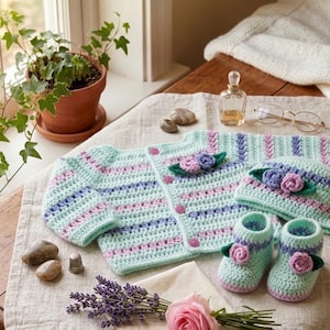 Peut inclure: Un ensemble crocheté à la main comprenant un gilet à manches longues, un bonnet assorti et une paire de chaussons. Les pièces sont réalisées en fil vert menthe clair avec des rayures horizontales lavande et rose, ornées de petites fleurs.
