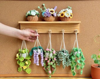 May include: Crocheted hanging plant decorations in various shades of green, purple, and pink, with white hanging cords. Three crocheted flower arrangements sit on a wooden shelf above. The background is a corkboard.
