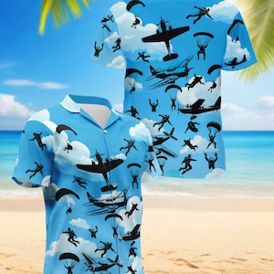 May include: Light blue short-sleeved shirt with a sky-themed design. The shirt features silhouettes of aeroplanes, parachutists, and clouds. The design is repeated on the back of the shirt. The shirt is displayed on a beach setting.