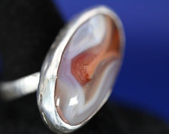 May include: A silver ring with an oval-shaped stone. The stone features a pattern of white, orange, and red hues. The ring's band is textured, and the setting is smooth. The ring is a piece of jewelry.