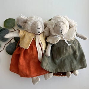 May include: Two handmade sheep dolls with cream-colored woolly heads and stitched facial features. One doll wears an orange dress and yellow vest, the other a green dress and white collar. The dolls are positioned on a woven base with green leaf accents.