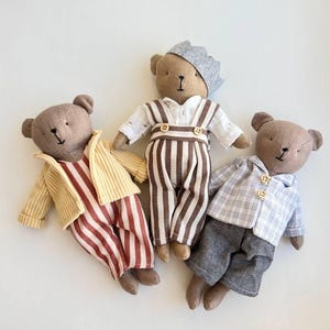 Könnte beinhalten: Drei handgefertigte Teddybären in Miniatur-Outfits. Einer trägt ein rot-weiß gestreiftes Outfit mit einer gelben Jacke. Ein anderer trägt eine braun-weiß gestreifte Latzhose und eine graue Krone. Der dritte trägt ein blau kariertes Hemd und graue Shorts.
