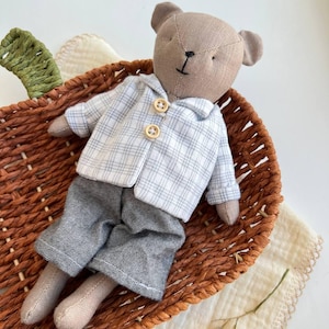 Könnte beinhalten: Eine handgefertigte Teddybär-Puppe mit beigem Körper, gekleidet in einer hellblau-weiß karierten Jacke und grauer Hose. Die Puppe liegt in einem braunen Weidenkorb. Das Outfit der Puppe hat Holzknöpfe. Der Korb ist mit einem cremefarbenen Tuch ausgekleidet.