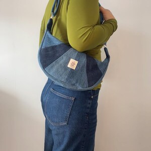Peut inclure: Un sac à bandoulière en forme de croissant fabriqué à partir de tissu en jean patchwork dans différentes nuances de bleu. Le sac présente une étiquette en tissu rectangulaire claire sur le devant avec un motif floral circulaire et du texte.