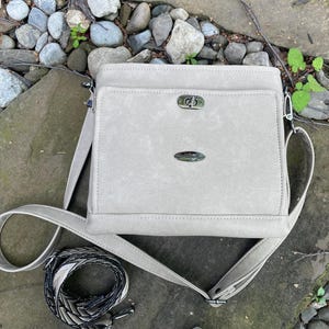 May include: A pale grey crossbody bag with a flap pocket and a detachable strap. The bag features silver-toned hardware and a decorative emblem. A patterned strap is coiled beside the bag.
