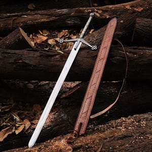 May include: A long, silver sword with an ornate hilt and crossguard lies next to a brown leather sheath. The sword's blade is polished. The sheath has a decorative pattern and a strap. The items are displayed on wooden logs.