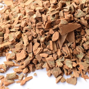 May include: A pile of small, irregular, dried cinnamon bark pieces. The fragments are reddish-brown in colour with a rough, woody texture, showing a mix of light and dark brown tones throughout the collection of bark chips.