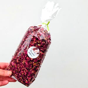 May include: A clear plastic bag filled with dried deep pink and burgundy rose petals. The bag is tied at the top with a thin green ribbon and features a circular white sticker on the front with floral graphics and the text Rose petal potpourri.