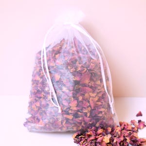 May include: A sheer white organza drawstring bag filled with dried rose petals in shades of deep pink, magenta, and orange. A small pile of the same dried rose petals is scattered on the surface in front of the bag, which is tied with white ribbons.