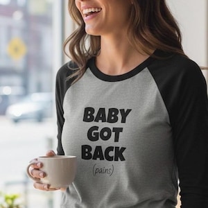 Könnte beinhalten: Graues Raglan-Shirt mit schwarzen Ärmeln und dem Text "BABY GOT BACK (pains)" in fetten schwarzen Buchstaben. Die Person hält eine beige Tasse.