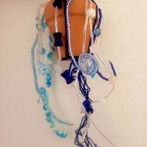 May include: A hanging terracotta pot with blue and white beaded embellishments. The pot is suspended by a blue macrame cord. Decorative strands of beads in shades of blue and white cascade from the pot, creating a whimsical, handcrafted look.