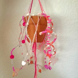 Könnte beinhalten: Ein hängender Terrakotta-Blumentopf, verziert mit rosa, weißen und orangefarbenen Perlen, Herzen und Schmetterlingsakzenten. Der Topf wird von einem rosa Makramee-Aufhänger gehalten und bildet einen dekorativen Pflanzenhalter. Das Design beinhaltet verschiedene Rosatöne.