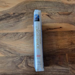 May include: Sealed VHS tape of "Lord of the Dance" by Michael Flatley. The spine is white with the title in gold and blue accents. The tape is in a clear plastic wrap.