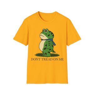 Puede incluir: Camiseta amarilla mostaza con un diseño de rana de dibujos animados. La rana verde tiene manchas y una bufanda azul. El texto "DON'T TREAD ON ME" está impreso debajo.