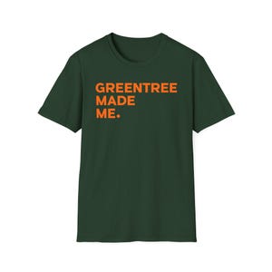 Puede incluir: Camiseta verde oscuro con el texto naranja "GREENTREE MADE ME." La camiseta de manga corta está hecha de un material suave y tiene cuello redondo. El texto está centrado en la parte delantera de la camiseta.