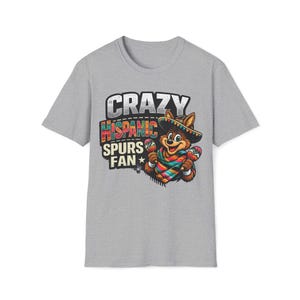 May include: A light grey t-shirt with a cartoon fox wearing a sombrero and holding maracas. The shirt features the text "CRAZY HISPANIC SPURS FAN" in a colourful, distressed font. The fox is wearing a colourful serape.