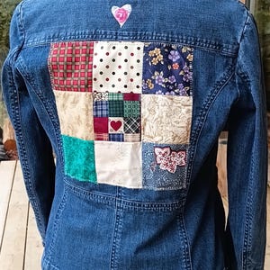 May include: A blue denim jacket with a patchwork design on the back. The patchwork features various fabric patterns, including floral, polka dots, and plaid. A small pink heart is stitched above the patchwork.