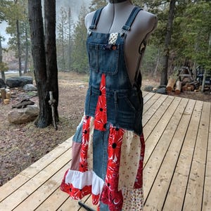Può includere: Salopette in denim con gonna patchwork con fiori rossi e tessuto fantasia. La salopette ha spalline regolabili e una tasca sul petto. La gonna ha un orlo a balze. L'indumento è esposto su un manichino all'aperto.
