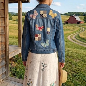 May include: A blue denim jacket featuring various fabric butterfly and square patches on the back, worn over a white eyelet midi skirt with floral embroidery and a ruffled hem. A straw hat is held in one hand, completing the ensemble.
