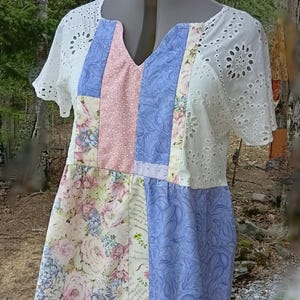Puede incluir: Vestido patchwork con escote en V, exhibido en un maniquí. El vestido presenta una combinación de telas, incluyendo mangas de encaje de ojal blanco, estampados florales y colores pastel lisos. El diseño general es una mezcla de texturas y patrones.