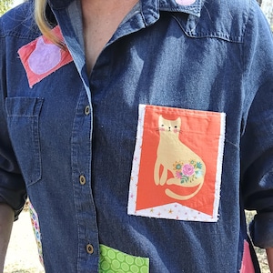 May include: A denim shirt with colorful patches and a cat graphic on the pocket. The shirt features a collared neckline, button-down front, and various fabric patches in pink, green, and patterned designs. The cat graphic is on an orange background.