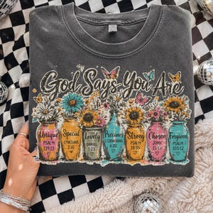 Pode incluir: Uma t-shirt de gola redonda cinzento-carvão com uma impressão gráfica de sete frascos de vidro coloridos cheios de flores. O texto diz God Says You Are seguido de palavras como Unique, Special, Lovely, Precious, Strong, Chosen e Forgiven com versículos.