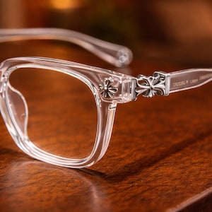 May include: Clear eyeglasses with a square frame design. The temples feature a decorative silver cross emblem. The glasses are set on a wooden surface, with a soft focus background.