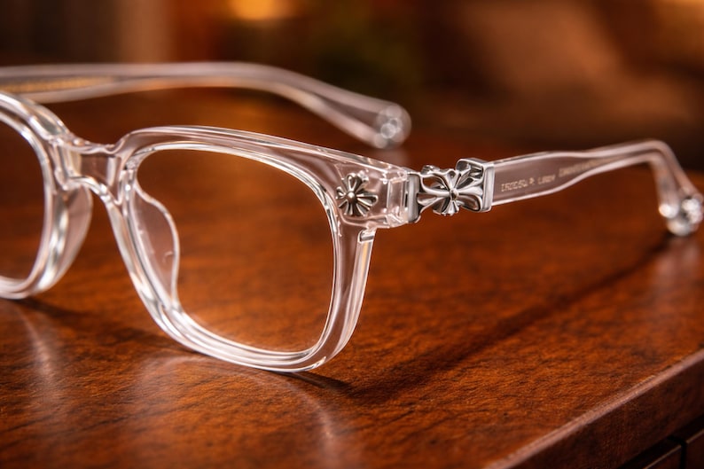 May include: Clear eyeglasses with a square frame design. The temples feature a decorative silver cross emblem. The glasses are set on a wooden surface, with a soft focus background.