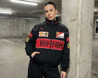 Peut inclure: Une veste zippée noire et rouge arborant le logo Ferrari sur la poitrine, accompagnée de divers écussons de sponsors dont Puma, Shell et UPS. La veste présente un col montant, des manches longues à poignets côtelés et divers emblèmes de course.