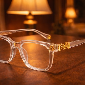 May include: Clear eyeglasses with a square frame and gold-colored decorative accents on the temples. The glasses are set against a warm, brown background, with a soft light illuminating the scene.