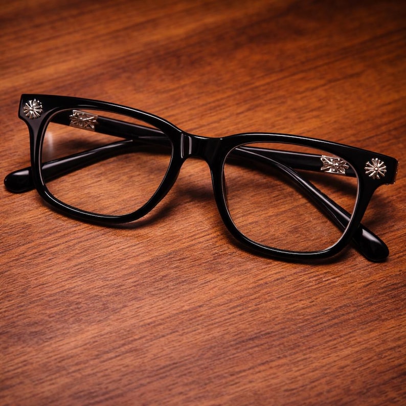 May include: Black eyeglasses with a classic rectangular frame design. The temples feature silver-colored decorative accents. The glasses are set against a warm, wooden surface, highlighting their sleek, modern aesthetic.