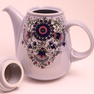 May include: A light blue ceramic teapot with a matching lid. The teapot features a floral design in shades of blue, pink, and green. The lid is off to the side. The teapot has a curved handle and a spout.