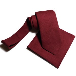 May include: A burgundy tie and pocket square set. The tie is made of a textured fabric and is folded in half. The pocket square is folded in a triangle shape.