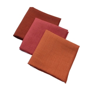 May include: Three linen napkins in shades of red, orange, and brown. The napkins are folded and arranged in a stack.