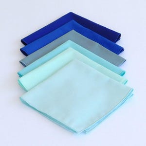 May include: A stack of six folded fabric napkins in shades of blue and turquoise. The napkins are arranged in a gradient from dark blue to light turquoise.