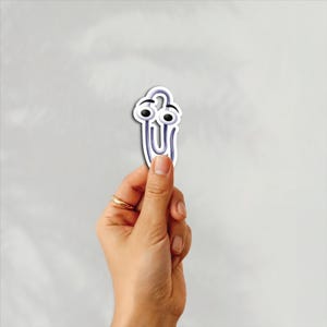 May include: A novelty paperclip sticker with a cartoon face, featuring large black eyes and a smiling expression. The sticker is held in a hand against a plain white background. The paperclip is purple and white.