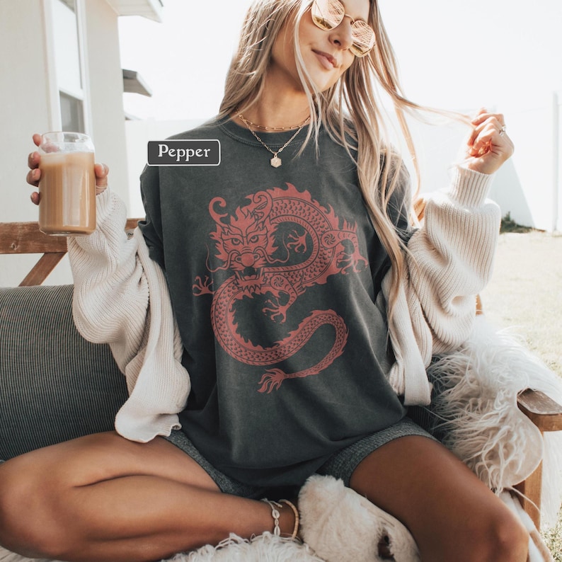 Puede incluir: Camiseta gris oscuro con un dise&ntilde;o de drag&oacute;n rojo. La palabra "Pepper" est&aacute; impresa en la camiseta. La camiseta se usa con una rebeca blanca y pantalones cortos grises. Se sostiene un vaso de caf&eacute; helado en la mano izquierda.