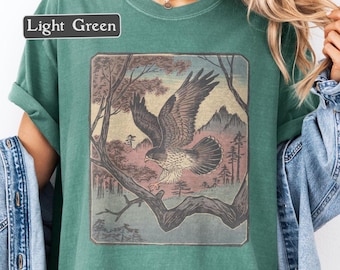 May include: Light green t-shirt featuring a framed illustration of a hawk in flight over a mountain landscape. The artwork is in muted tones of brown, gray, and pink. The text "Light Green" is in a black rectangle in the upper left.