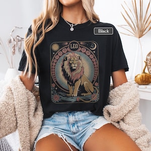 May include: Black t-shirt featuring a detailed illustration of a lion with the word "LEO" above it. The design includes a sun, moon, and floral elements. The shirt is labeled "Black".