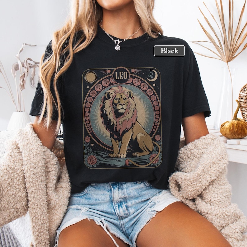 May include: Black t-shirt featuring a detailed illustration of a lion with the word "LEO" above it. The design includes a sun, moon, and floral elements. The shirt is labeled "Black".