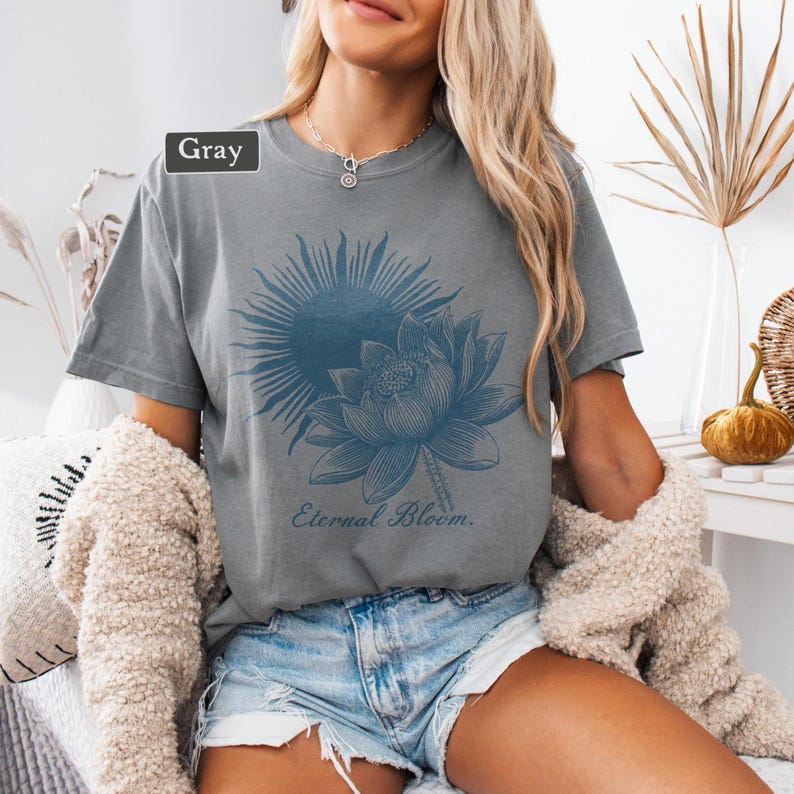 May include: Gray t-shirt featuring a blue sun and lotus flower graphic. The text "Eternal Bloom" is printed below the floral design. The shirt is made of a soft material.