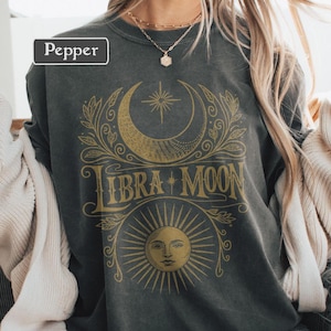 May include: Dark grey sweatshirt with a gold Libra Moon graphic. The design features a crescent moon, star, and sun with the words "Libra Moon". The word "Pepper" is in a white box in the upper left corner.