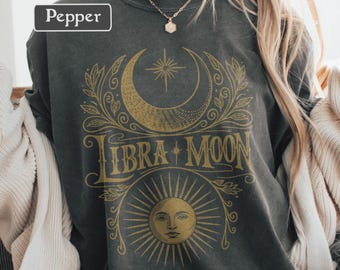 May include: Dark gray sweatshirt with a gold Libra Moon graphic. The design features a crescent moon, star, and sun with the words "Libra Moon". The word "Pepper" is in a white box in the upper left corner.