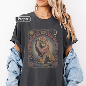 May include: Dark gray t-shirt featuring a detailed illustration of a lion, the zodiac sign Leo, and floral and celestial elements. The word "Pepper" is in a white rectangle in the upper left corner. The design is in shades of brown, pink, and blue.