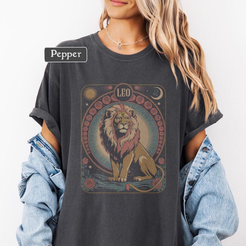May include: Dark gray t-shirt featuring a detailed illustration of a lion, the zodiac sign Leo, and floral and celestial elements. The word "Pepper" is in a white rectangle in the upper left corner. The design is in shades of brown, pink, and blue.