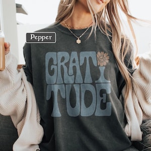 May include: Dark gray sweatshirt with the word "GRATITUDE" in blue, with a smiling sunflower graphic. The word "Pepper" is in a white box in the upper left corner. The sweatshirt is worn by a person with long blonde hair.