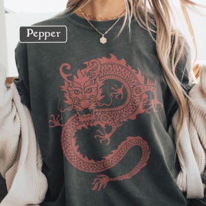 Puede incluir: Sudadera gris oscuro con un dise&ntilde;o de drag&oacute;n rojo. La palabra "Pepper" est&aacute; en un rect&aacute;ngulo blanco. El dise&ntilde;o del drag&oacute;n es detallado y cubre una gran parte de la parte delantera de la prenda.