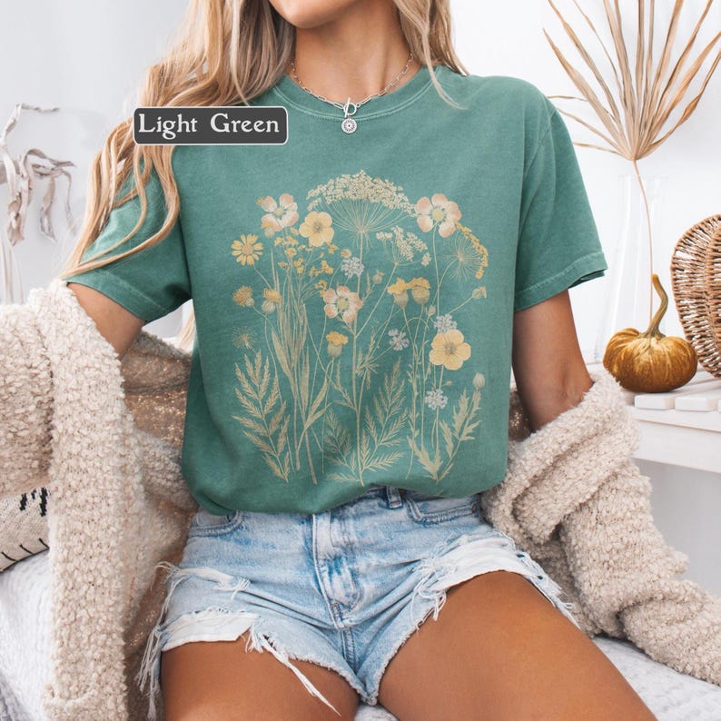 以下が含まれることがあります： 黄色とクリーム色の花柄が特徴のライトグリーンのTシャツ。Tシャツは半袖とクルーネックです。「Light Green」の文字が左上の白い長方形の中にあります。