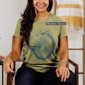 May include: Heather green t-shirt with rolled sleeves. The shirt features a blue graphic of a tree cross-section and the quote, "Adopt the pace of nature, her secret is patience." The shirt is worn by a person sitting in a wooden chair.